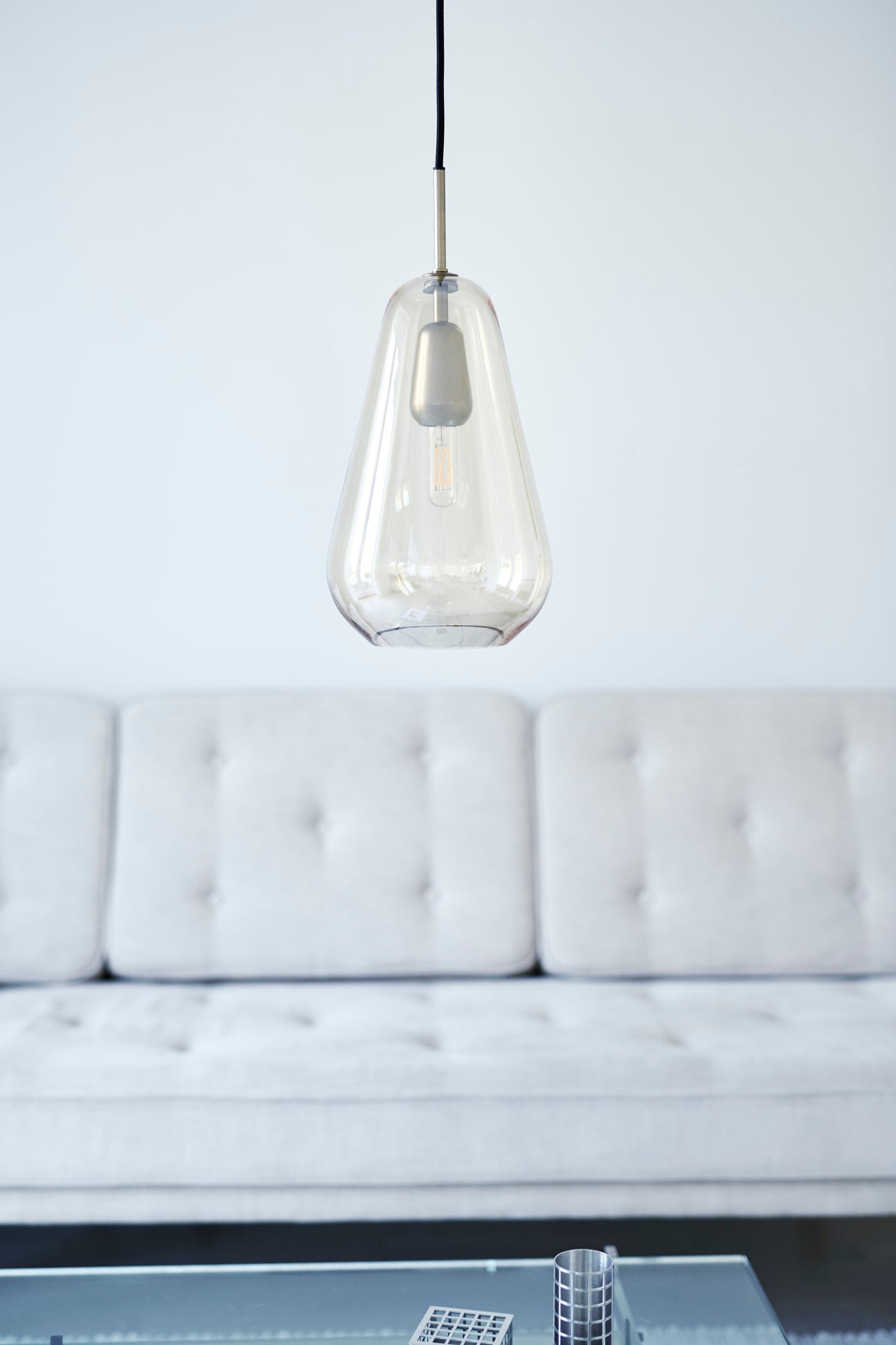 Dråbeformet pendel af gyldent glas med gyldent ophæng og sort ledning, med lys sofa i baggrunden