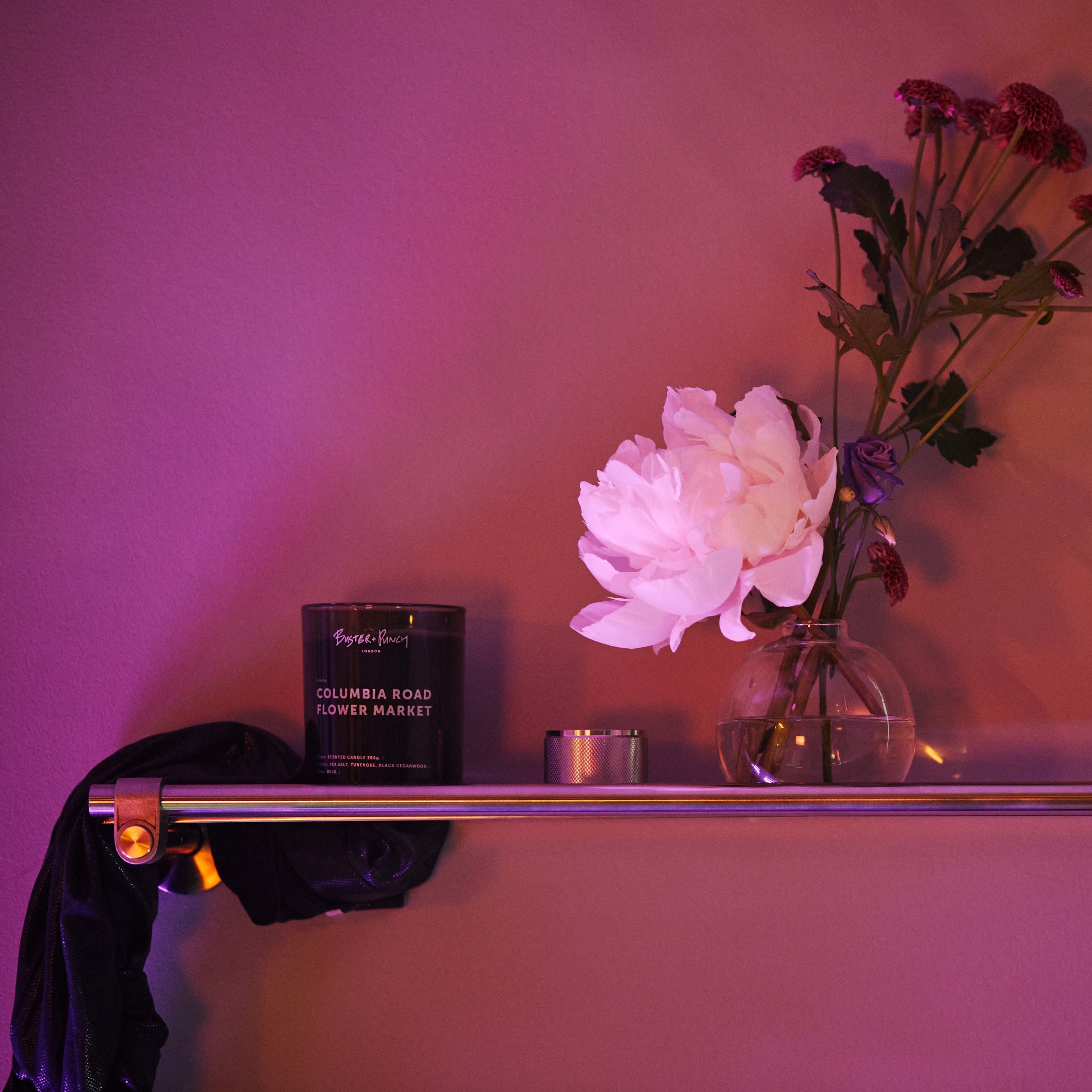 Duftlys af sort voks i røgfarvet glas med tændt flamme, på messinghylde i badeværelsesmiljø.