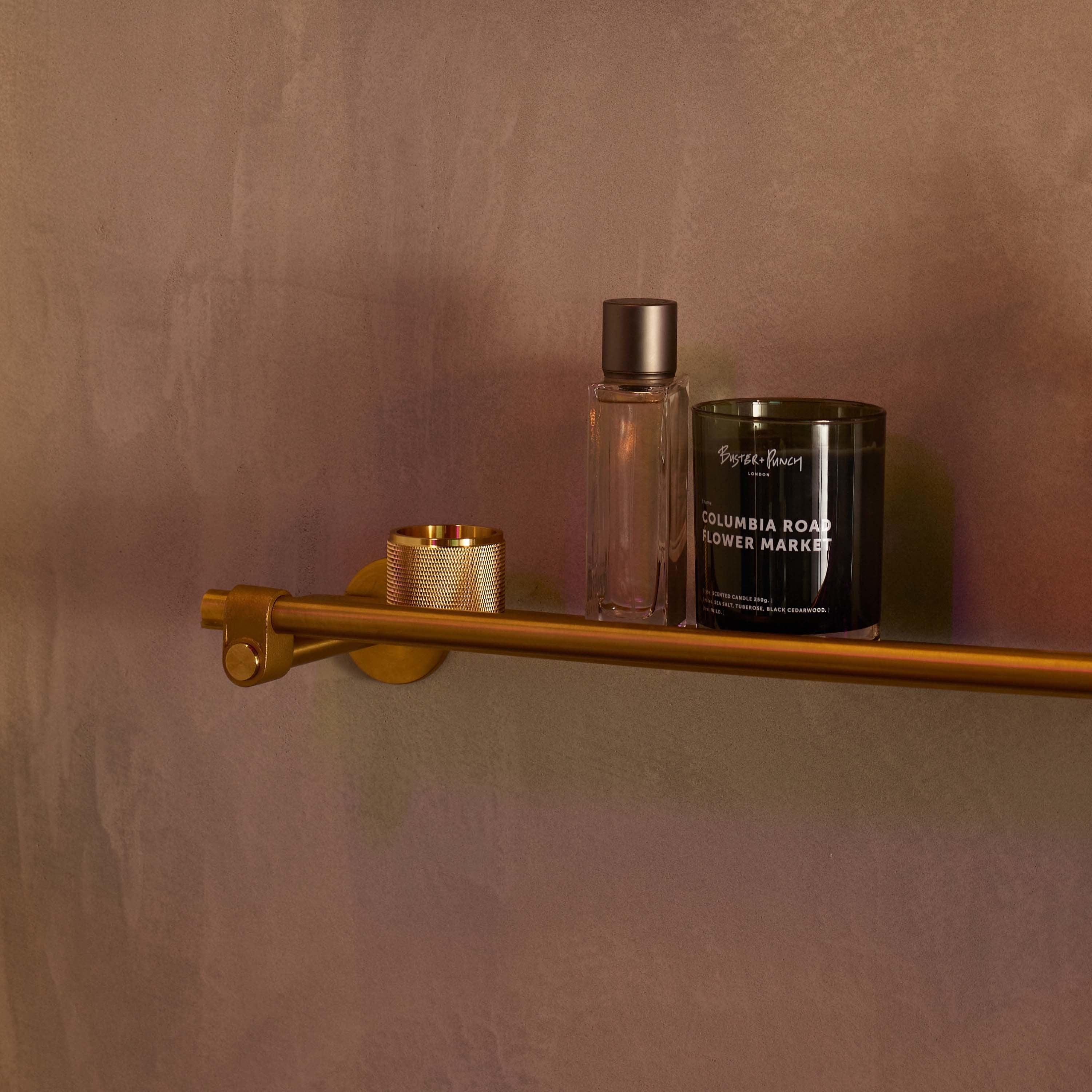 Duftlys af sort voks i røgfarvet glas med tændt flamme, på messinghylde i badeværelsesmiljø.