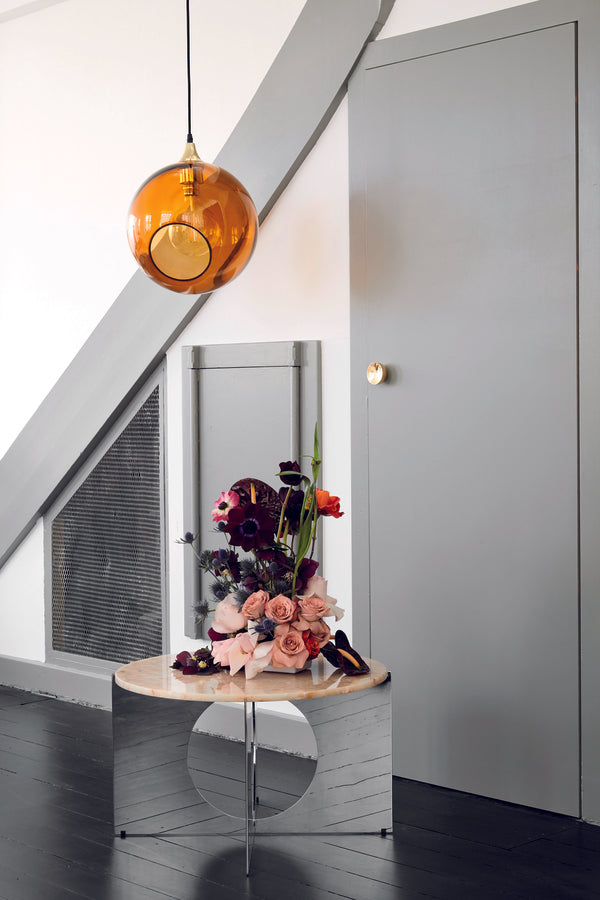 Rund lampe i orange/gult glas og med et afskåret stykke med guldkant og kig til pæren. Fatningen og rosetten er guldfarvet og ledningen er af sort stof.