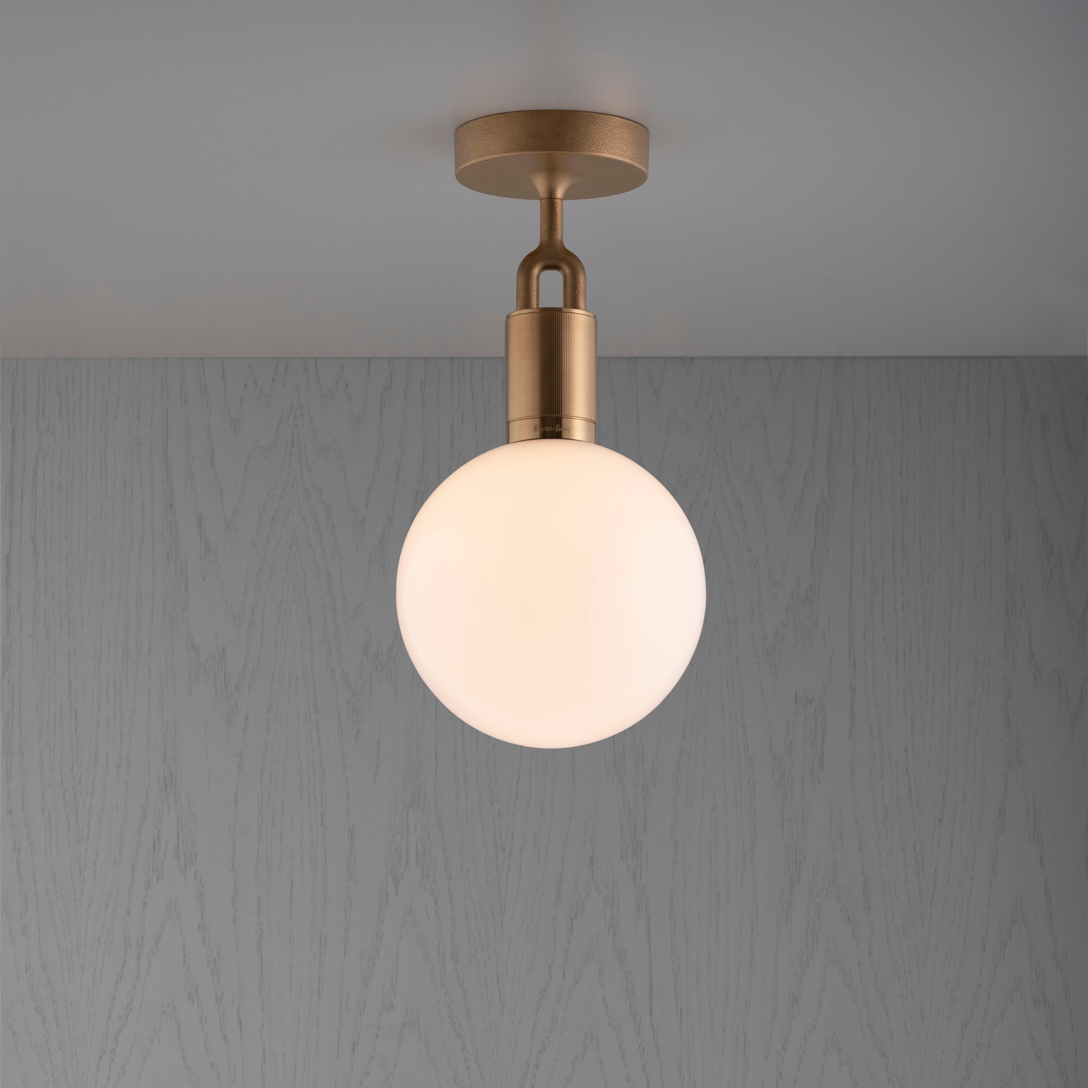 Loftlampe med rund skærm af opalglas og med fatning i messing, på baggrund af gråt træ.