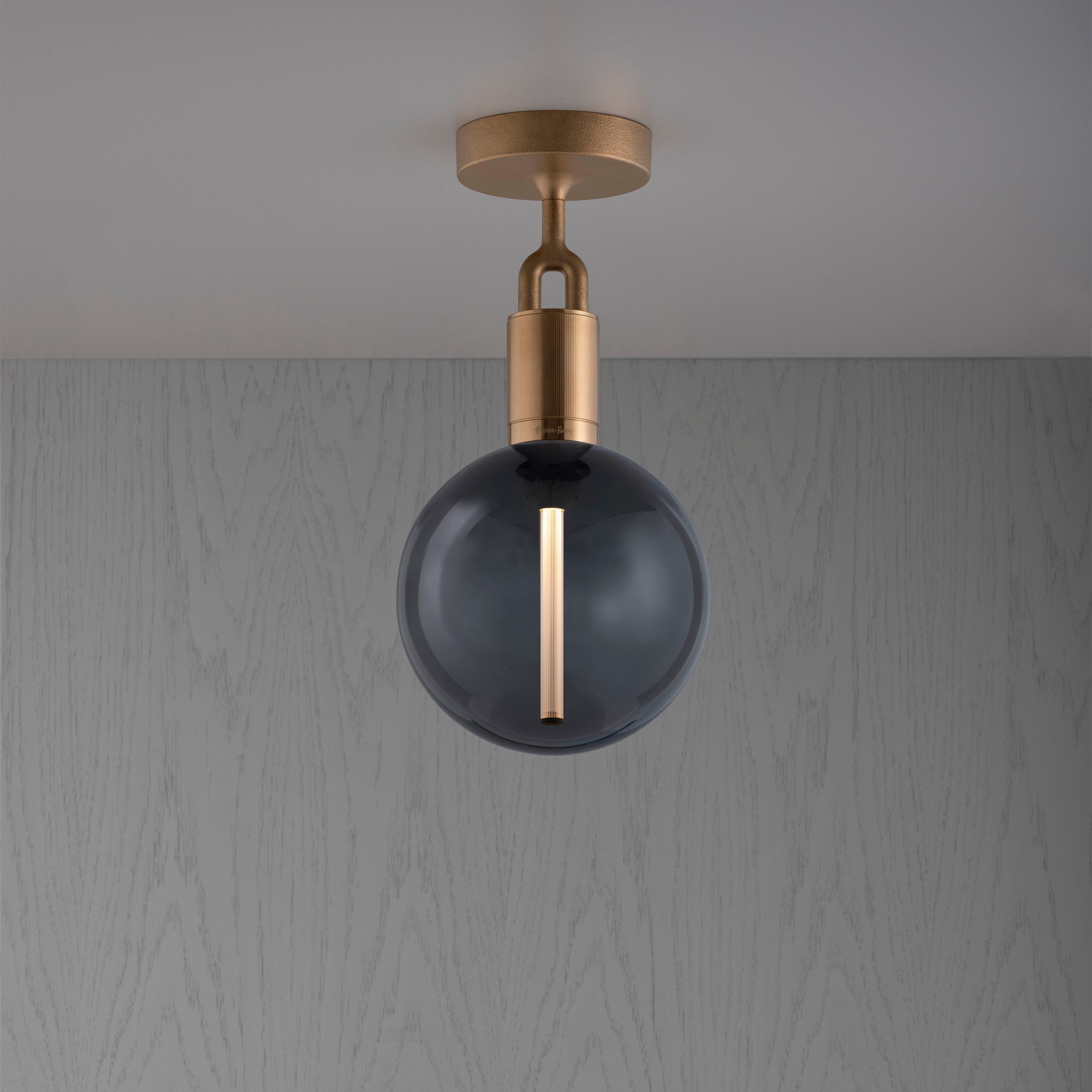 Loftlampe med rund skærm af røgfarvet glas og fatning i messing, på baggrund af grå trævæg.