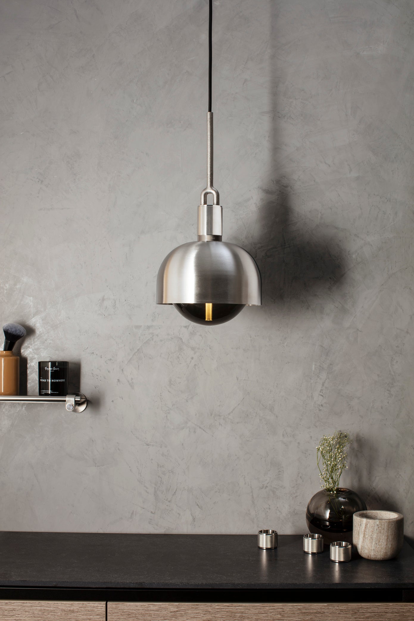 Pendel med fatning og skærm af stål samt pære i røgfarvet glas, hængende over køkkenbord.