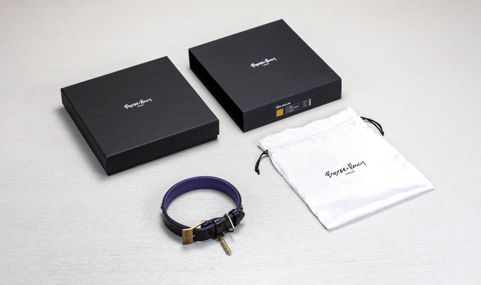 Æske indeholdende hundehalsbånd i sort og lilla læder med detaljer i messing, på hvid baggrund.