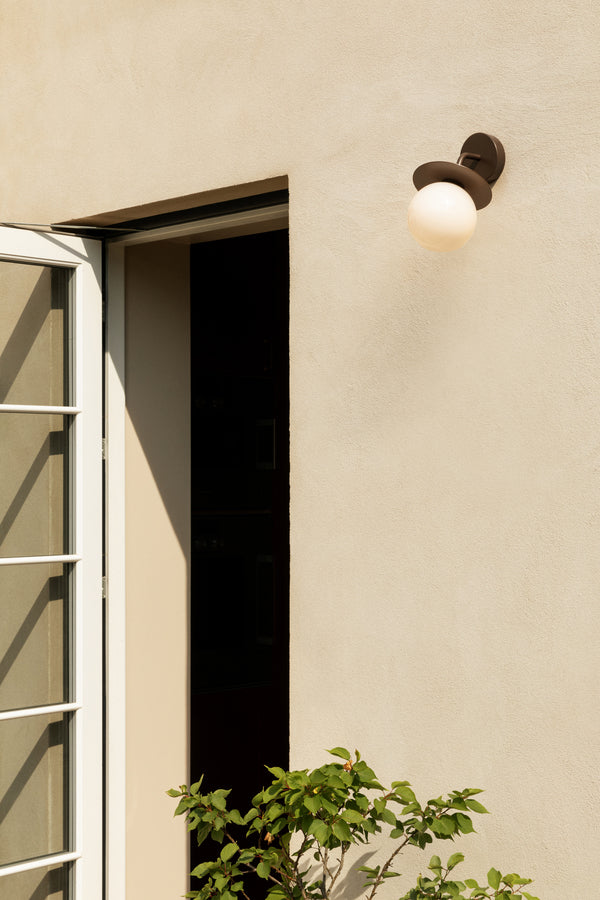 Væglampe med rund skærm af opalglas og bronzefarvet stel, monteret på lys mur ved siden af dør