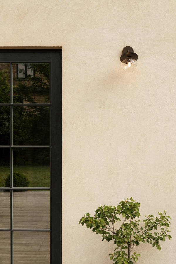 Væglampe med rund skærm af klart optikglas og bronzefarvet stel, på lys mur ved siden af dør