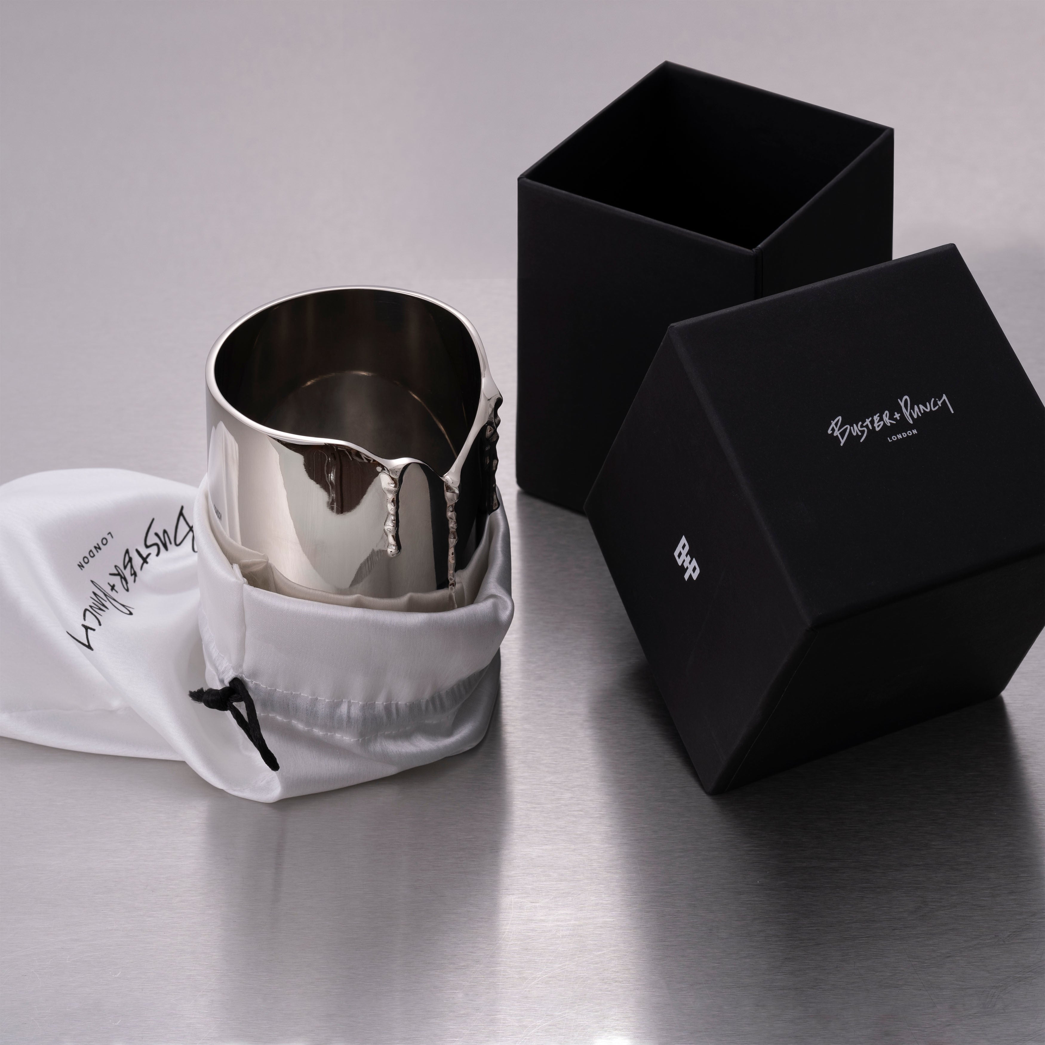 Æske indeholdende lysestage til duftlys i stål, på grå baggrund.