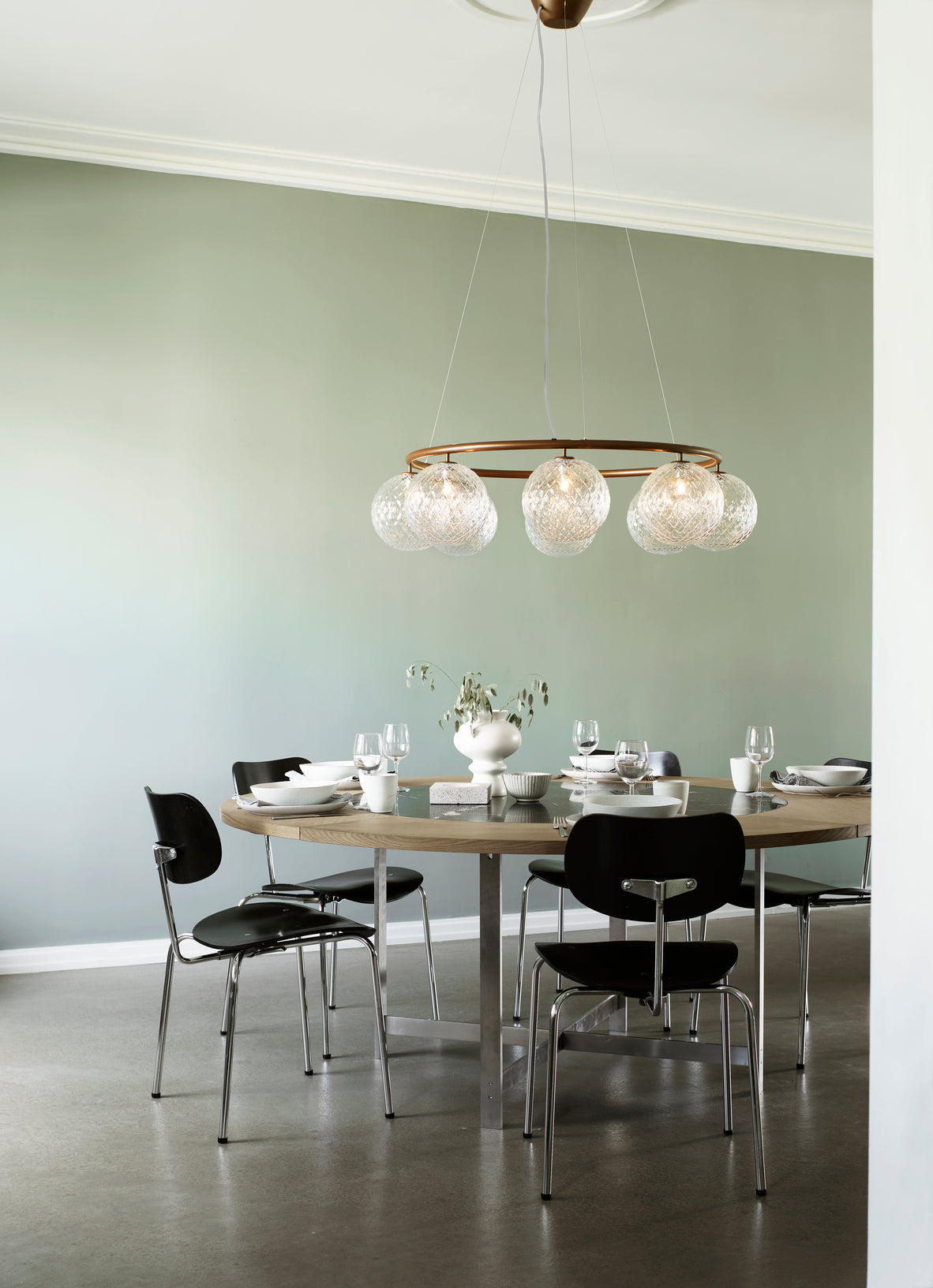 Lysekrone bestående af otte runde skærme i klart optikglas samt bruneret krone, hængende over spisebord
