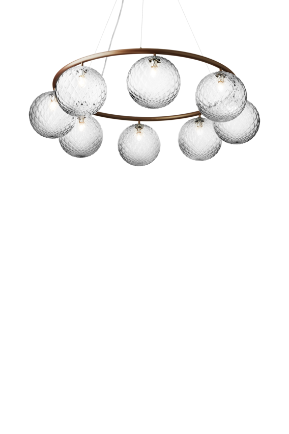 Lysekrone bestående af otte runde skærme i klart optikglas samt bruneret krone, på hvid baggrund