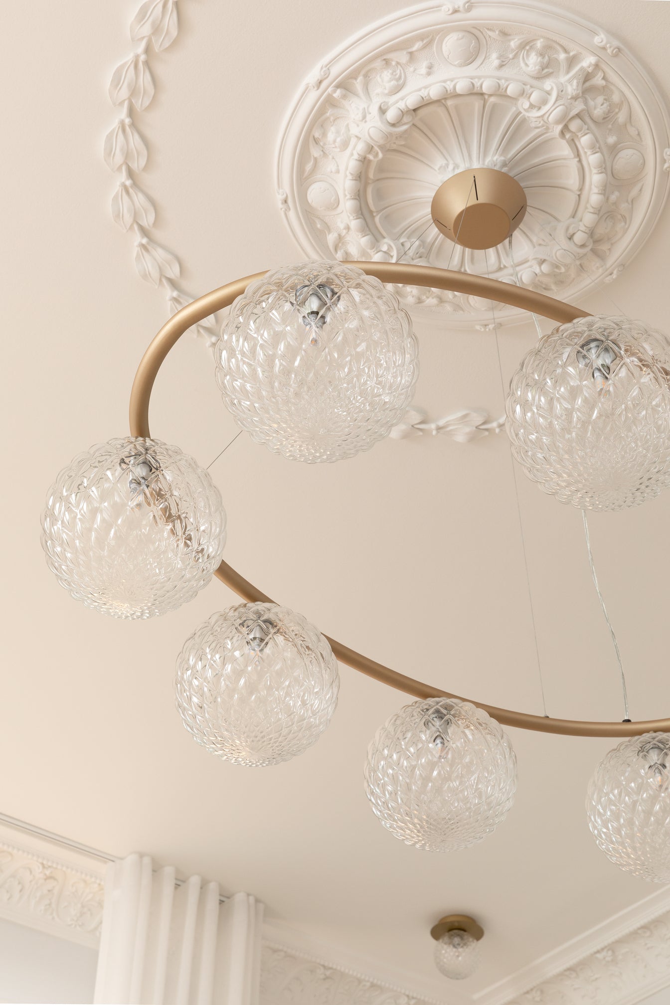 Lysekrone bestående af otte runde skærme i klart optikglas samt messingfarvet krone, hængende i stuk roset i loftet