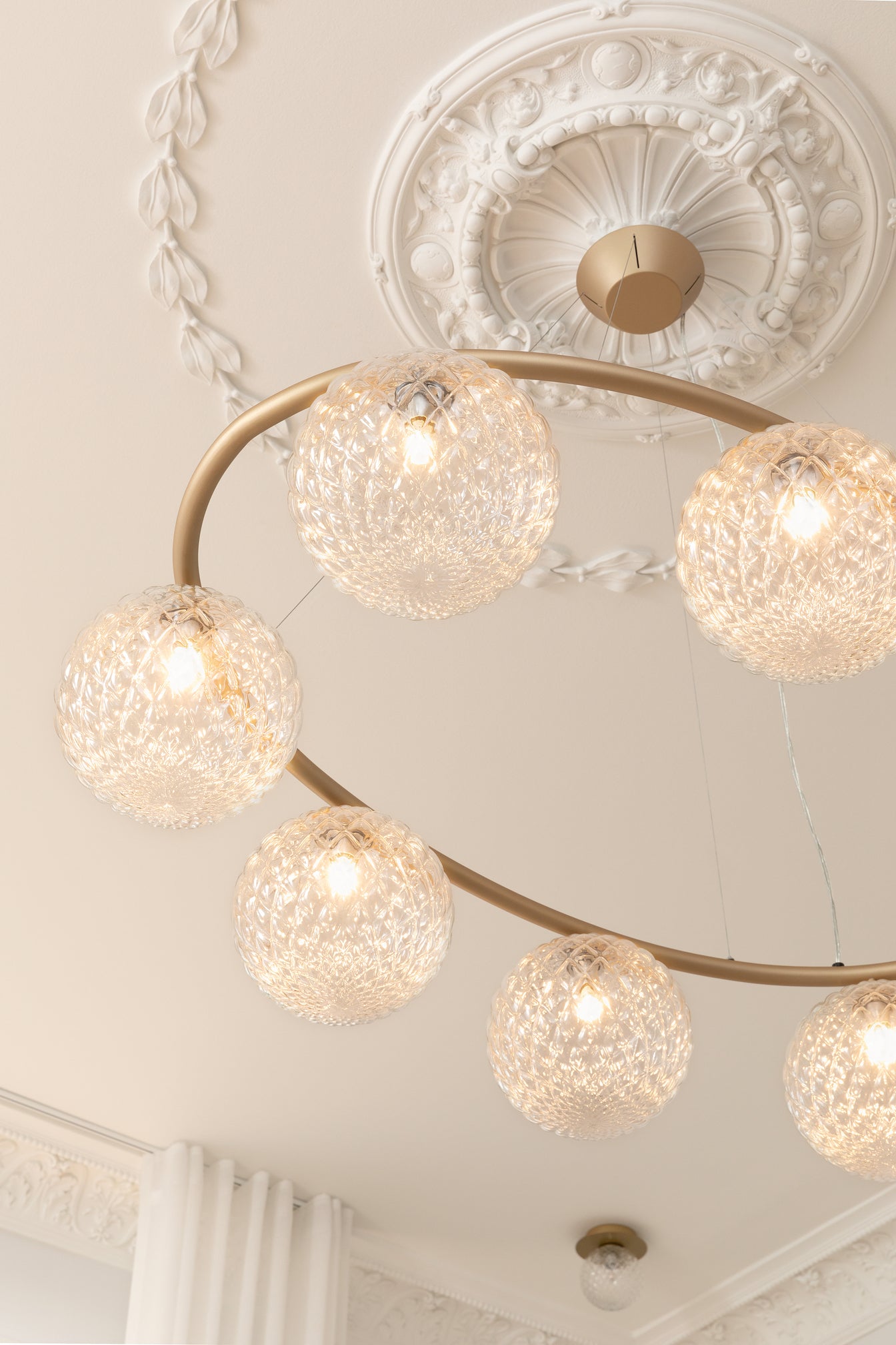 Lysekrone bestående af otte runde skærme i klart optikglas samt messingfarvet krone, hængende i stuk roset i loftet