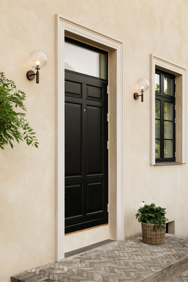 Væglamper med runde skærme af klart optikglas og bruneret stel, ved siden af hoveddør på gul mur