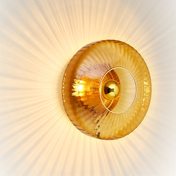 Rund væglampe i amber (orange/gul) plisseret glas, som skaber et lysspil på væggen.