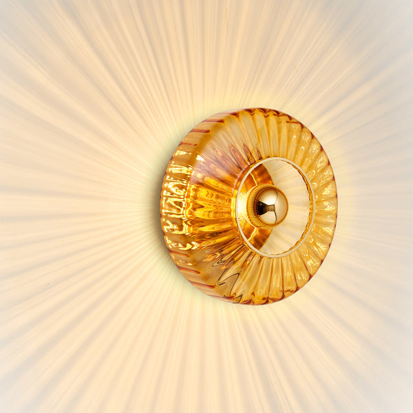 Rund væglampe i amber (orange/gul) plisseret glas, som skaber et lysspil på væggen.
