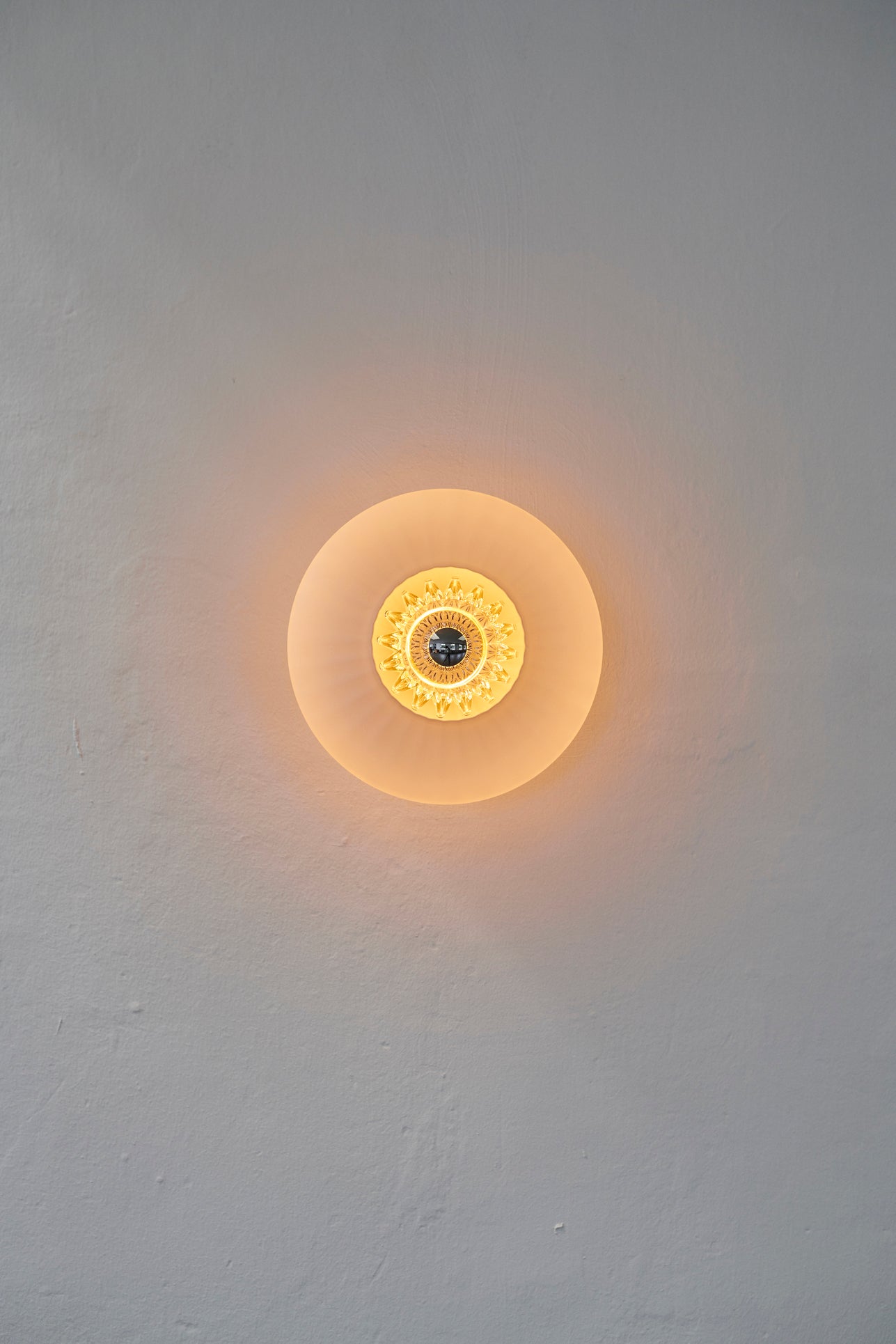 Rund væglampe i opal plisseret glas, som skaber et lysspil på væggen.