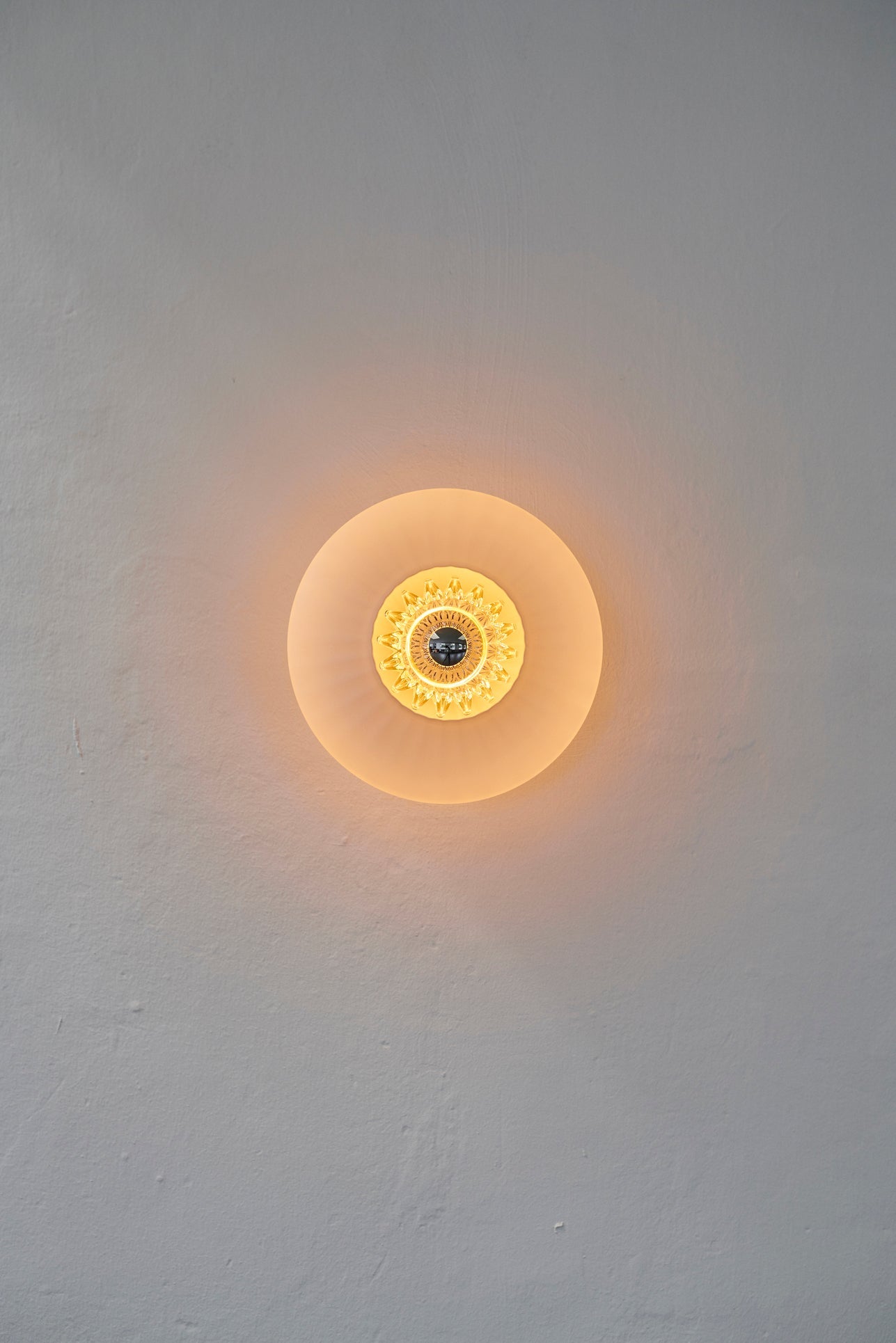 Rund væglampe i amber plisseret glas, som skaber et lysspil på væggen.