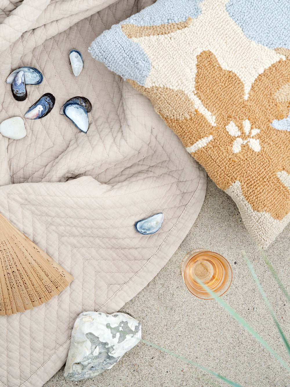 Beige tæppe ligger på sand med muslinger og forskelligfarvede pyntepuder.