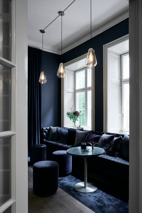 Tre dråbeformede pendler af gyldent glas med gyldent ophæng og sort ledning, hængende over blå sofa og foran blåmalede vægge.