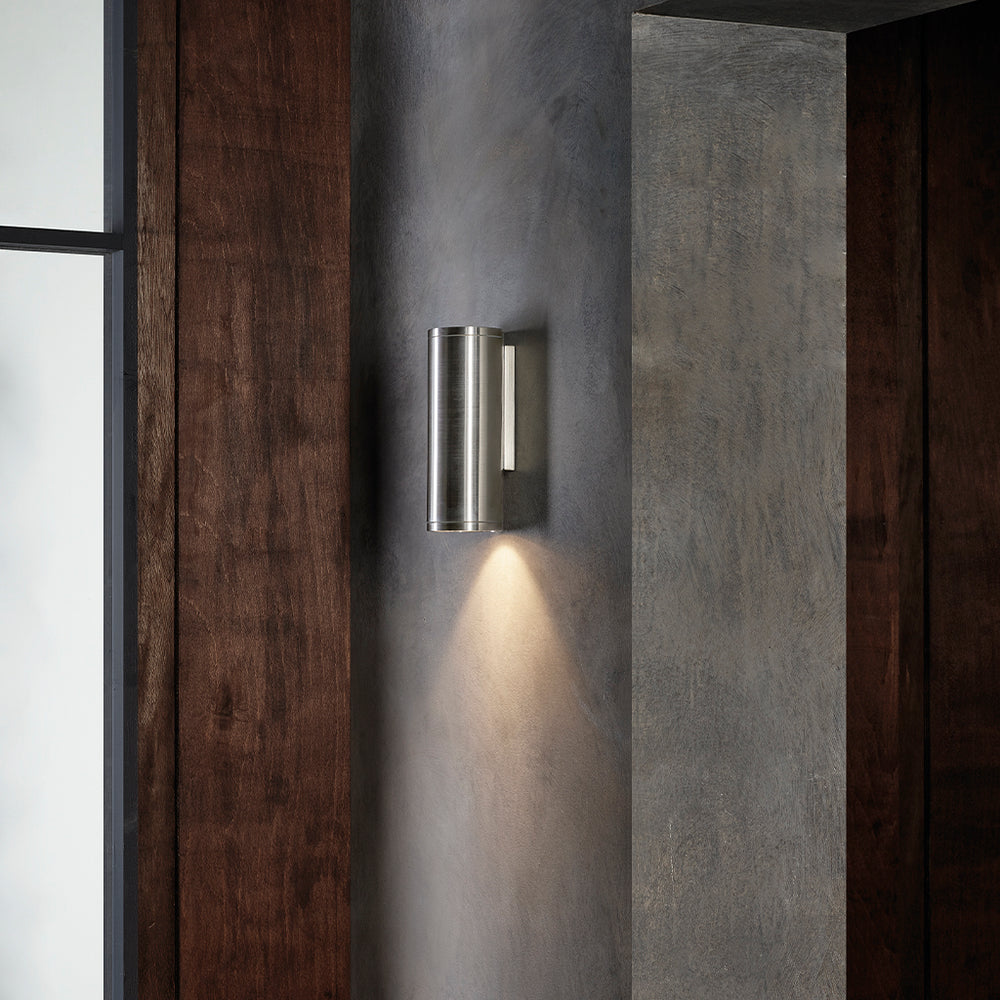 Udendørslampe i rustfrit stål med et rundt og aflangt design. Lampen har nedadgående kegleformet lys.