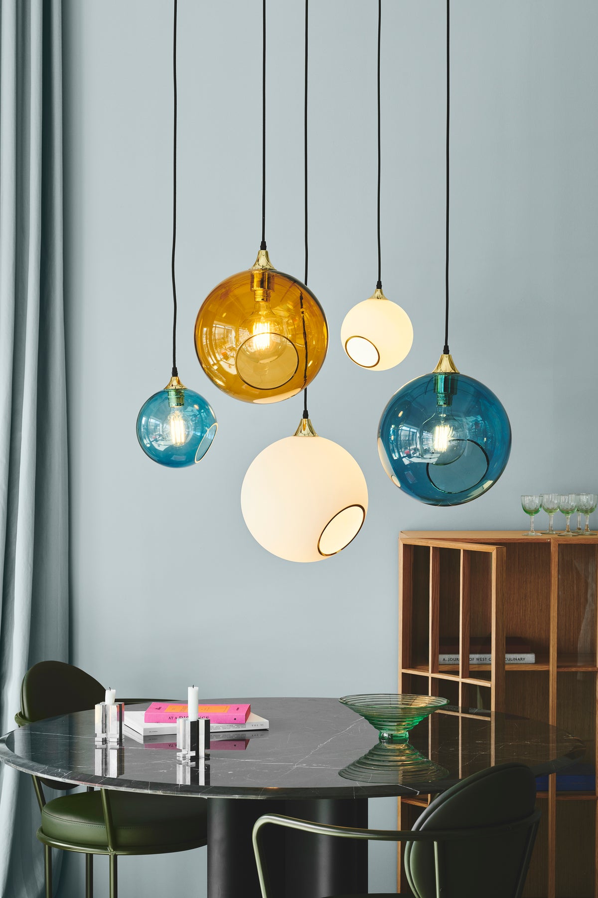 Rund lampe i blåt glas og med et afskåret stykke med guldkant og kig til pæren. Fatningen og rosetten er guldfarvet og ledningen er af sort stof.