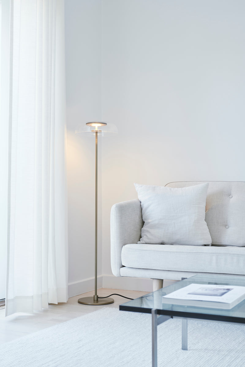 Gulvlampe med gyldent stel og fatning samt klar glasskærm, i dagligstuemiljø