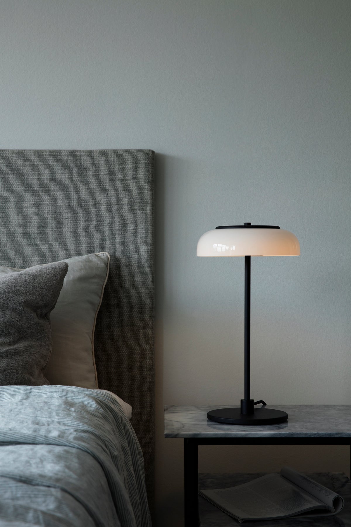 Bordlampe med sort stel og fatning og glasskærm i opalglas, i sengebord ved siden af seng