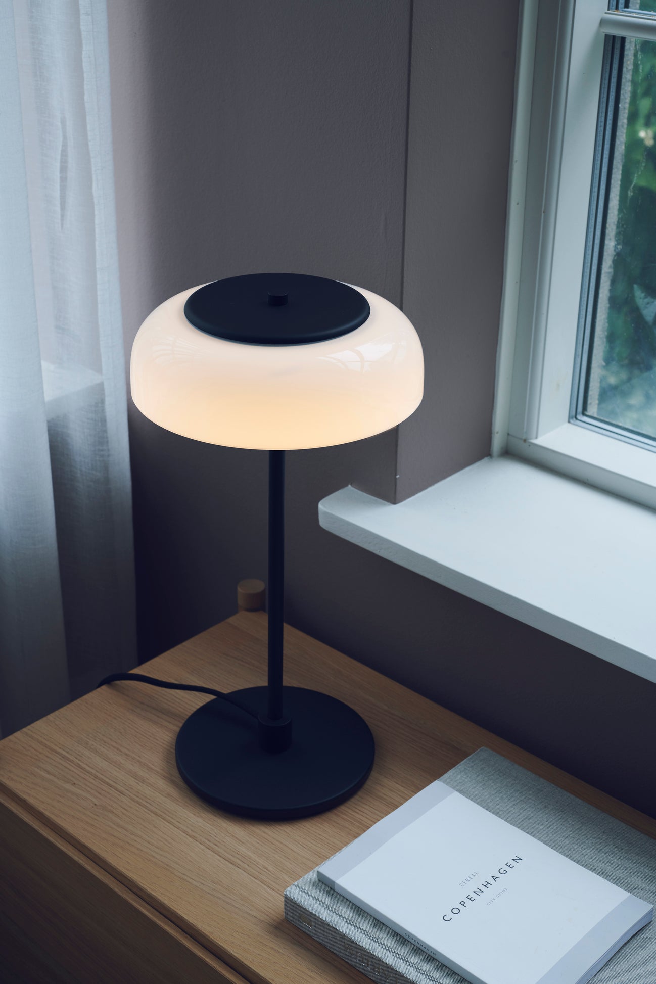 Bordlampe med sort stel og fatning og glasskærm i opalglas, i dagligstuemiljø