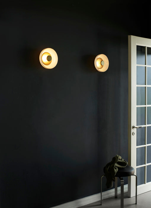 Rund væglampe af opalglas med gylden fatning, i dagligstuemiljø på mørk væg