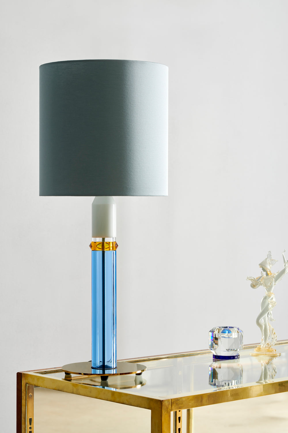 Bordlampe med fod i blåt, hvidt og ravfarvet krystal, med grå lampeskærm, stående på glasreol.