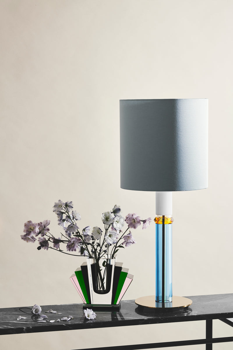Bordlampe med fod i blåt, hvidt og ravfarvet krystal, med grå lampeskærm samt krystalvase med blomster, stående på marmor konsolbord.
