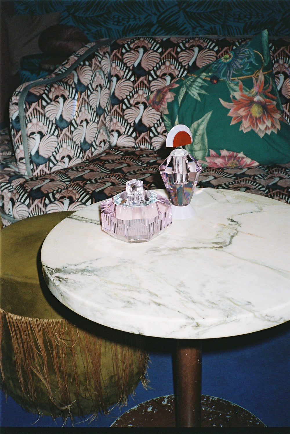 Opstilling af duftlys i farvet krystalbeholder samt flakon i farvet krystal, stående på rundt marmorbord med loungemøbler i baggrunden.