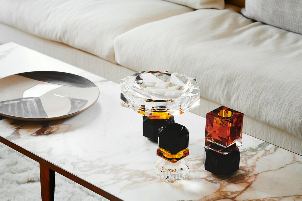 Opstilling af lysestager og skål i farvet krystal, på sofabord i marmor med sofa i baggrunden.