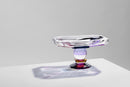 Aflang skål på fod i klar, lilla og bourdeaux krystal, på hvidt bord og lysegrå baggrund.