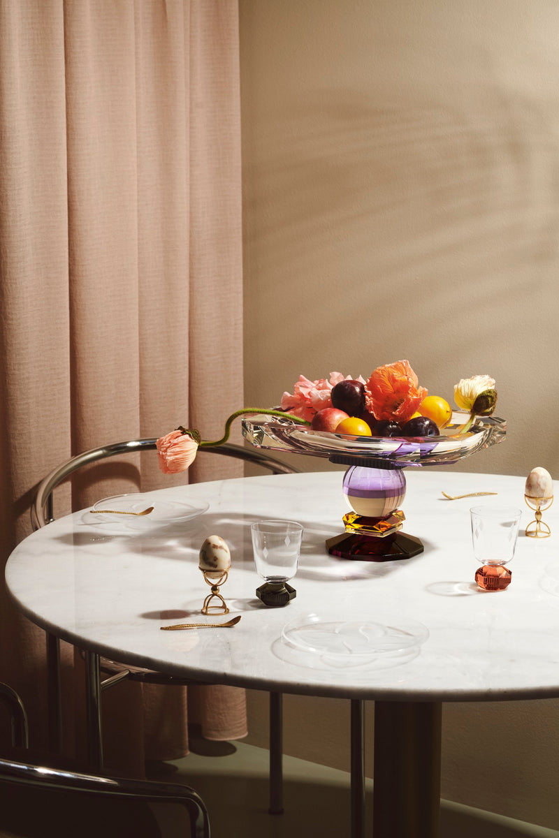 Aflang skål på fod i klar, lilla og bourdeaux krystal med frugt og blomster i, på hvidt bord og med beige gardin i baggrund.