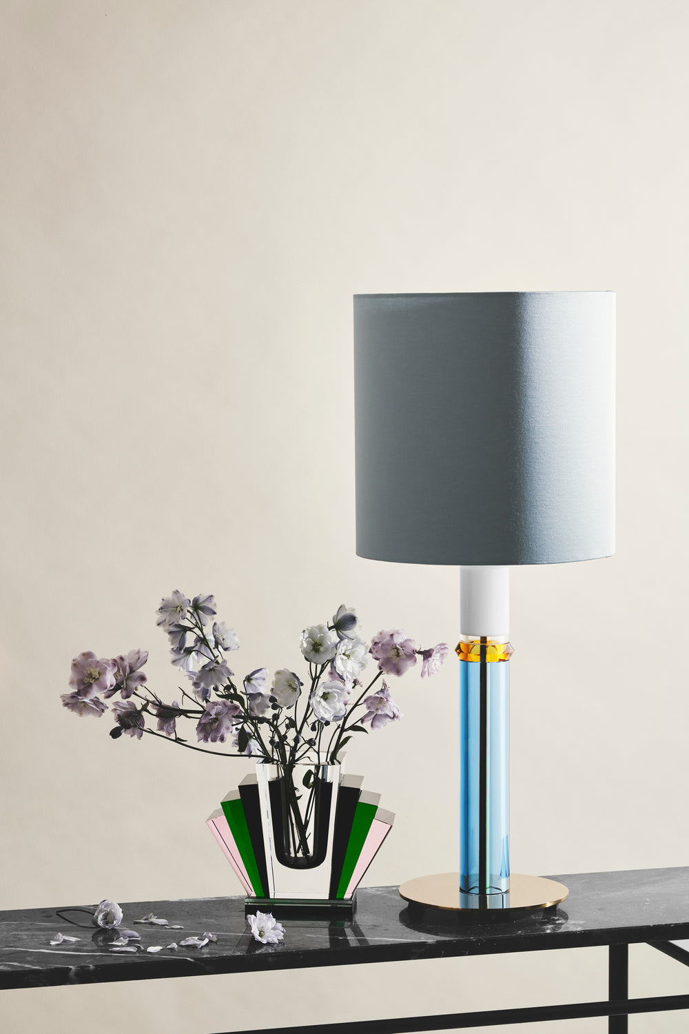 Opsætning af vase i klar, sort, grøn og lyserød krystal med blomster i samt bordlampe stående på marmorbord og hvid baggrund.