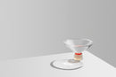 Kegleformet skål med fod i klar, hvid og rød krystal, på hvidt bord med lysegrå baggrund.