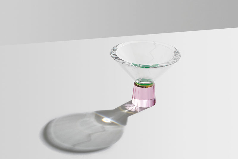 Kegleformet vase på fod i klar, lyserød og grøn krystal, på hvidt bord og lysegrå baggrund.