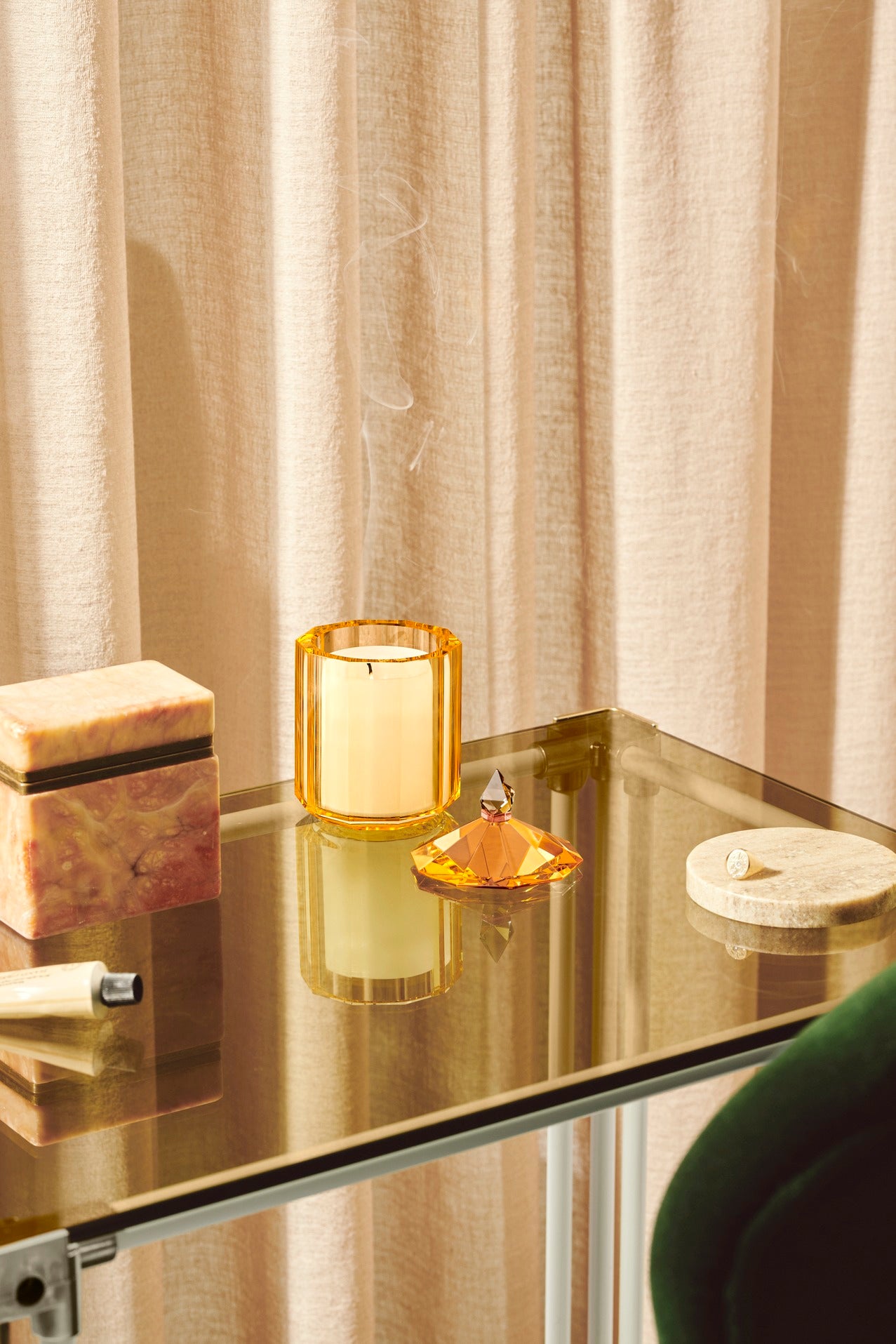 Duftlys i beholder af gul krystal med aftraget låg, på glasbord og med beige gardin i baggrunden