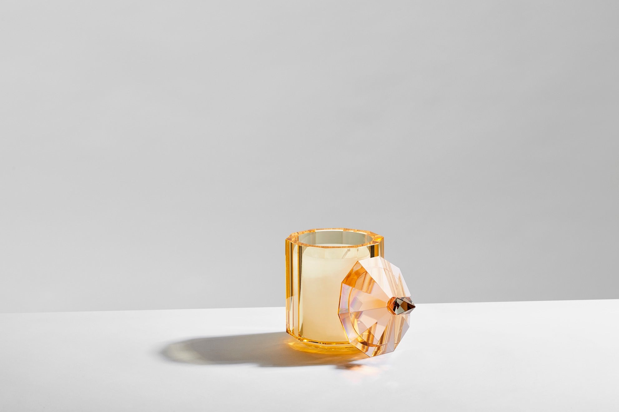 Duftlys i beholder af gul krystal med aftaget låg, på hvidt bord og med grå baggrund