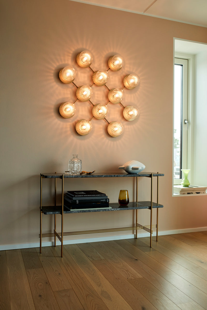 Væglampe med lampeskærme af klart optikglas og gylden fatning og stel, på beige væg i dagligstuemiljø