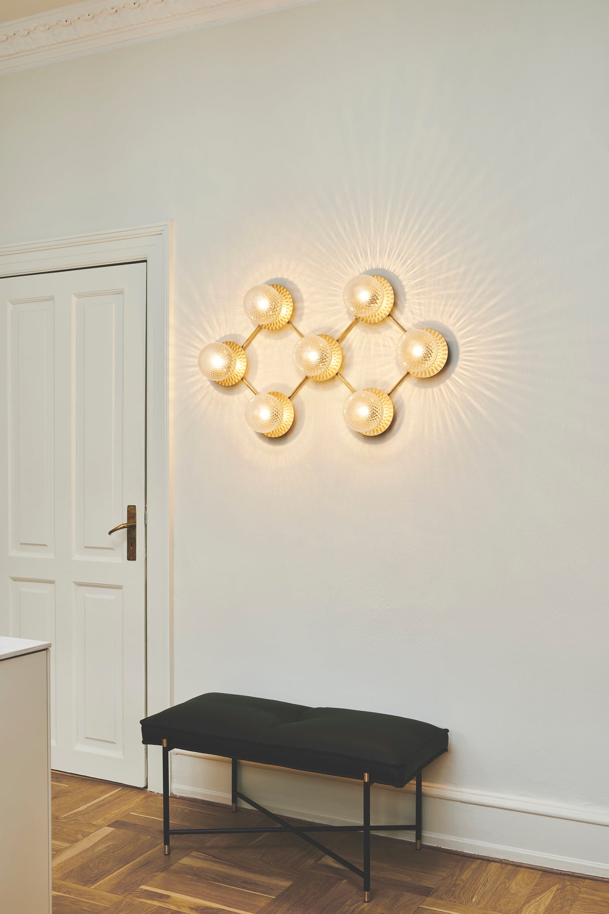 Væglampe med lampeskærme af klart optikglas og gylden fatning og stel, på hvid væg over bænk