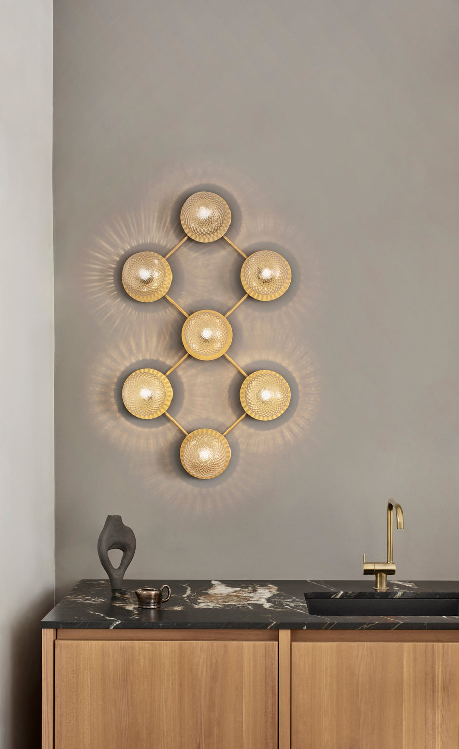 Væglampe med lampeskærme af klart optikglas og gylden fatning og stel, hængende på væg ved køkkenbord