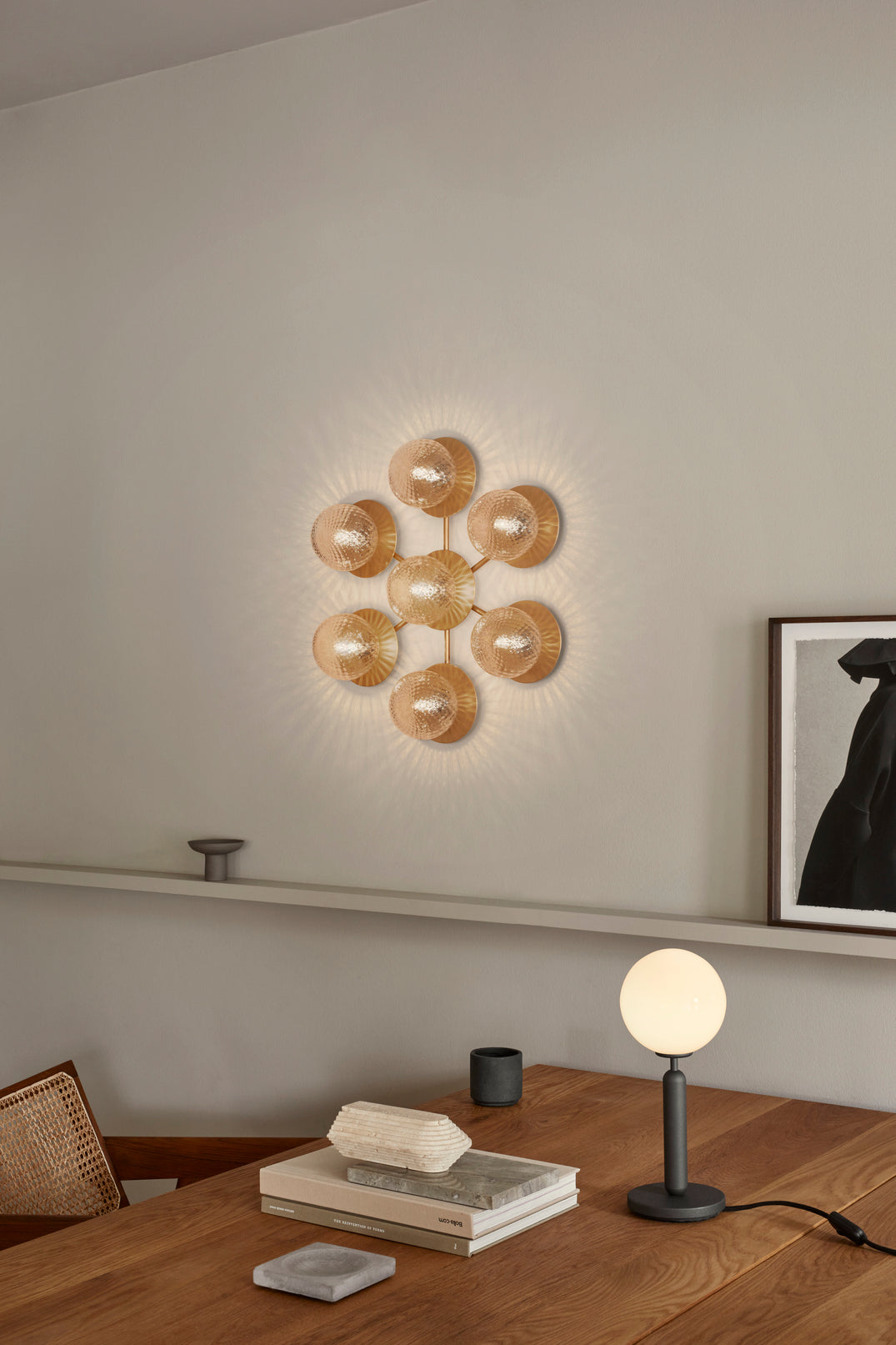 Væglampe med lampeskærme af klart optikglas og gylden fatning og stel, på hvid væg ved siden af skrivebord