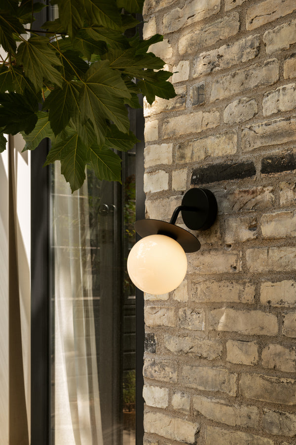 Væglampe med rund skærm af opalglas og sort stel, monteret på gul murstensvæg ved siden af terrassedør
