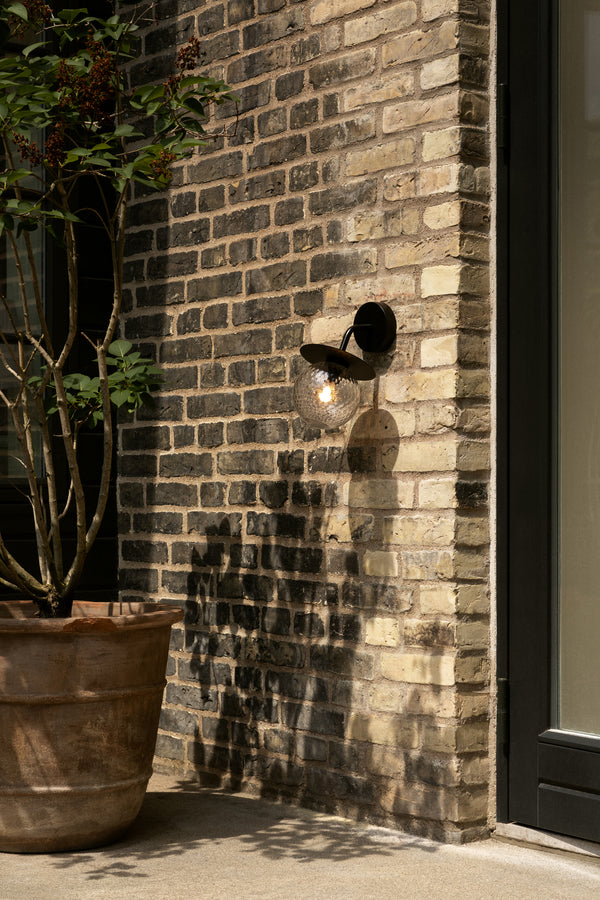 Væglampe med rund skærm af klart optikglas og sort stel, monteret på gul murstensvæg ved siden af terrassedør