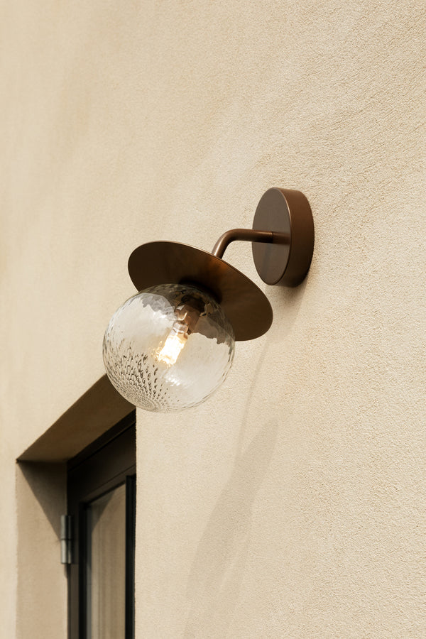 Væglampe med rund skærm af klart optikglas og bronzefarvet stel, på lys mur