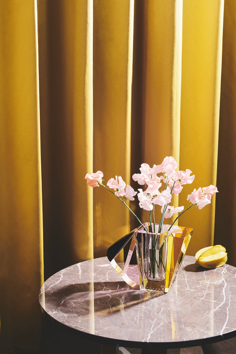 Vase i klar, brun, gul og lyserød krystal med blomster i, på rundt marmorbord på baggrund af gult gardin.