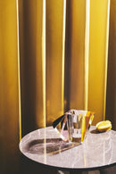 Vase i klar, brun, gul og lyserød krystal, på rundt marmorbord på baggrund af gult gardin.