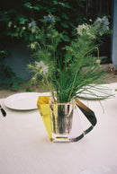 Vase i klar, brun, gul og lyserød krystal med blomster i, på bord med hvidt dug og beplantning i baggrunden.