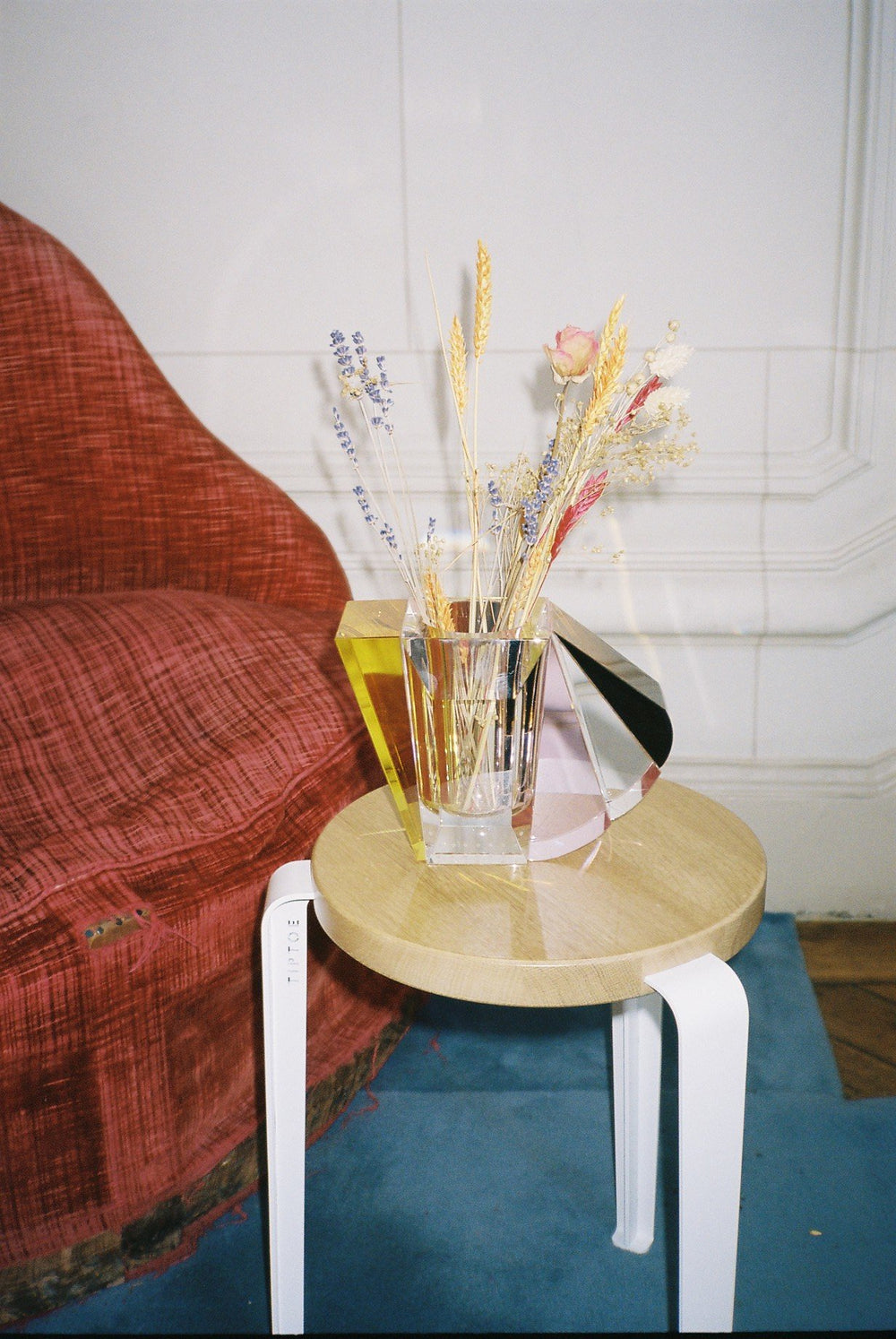 Vase i klar, brun, gul og lyserød krystal med blomster i, på træskammel ved siden af en rød lænestol.